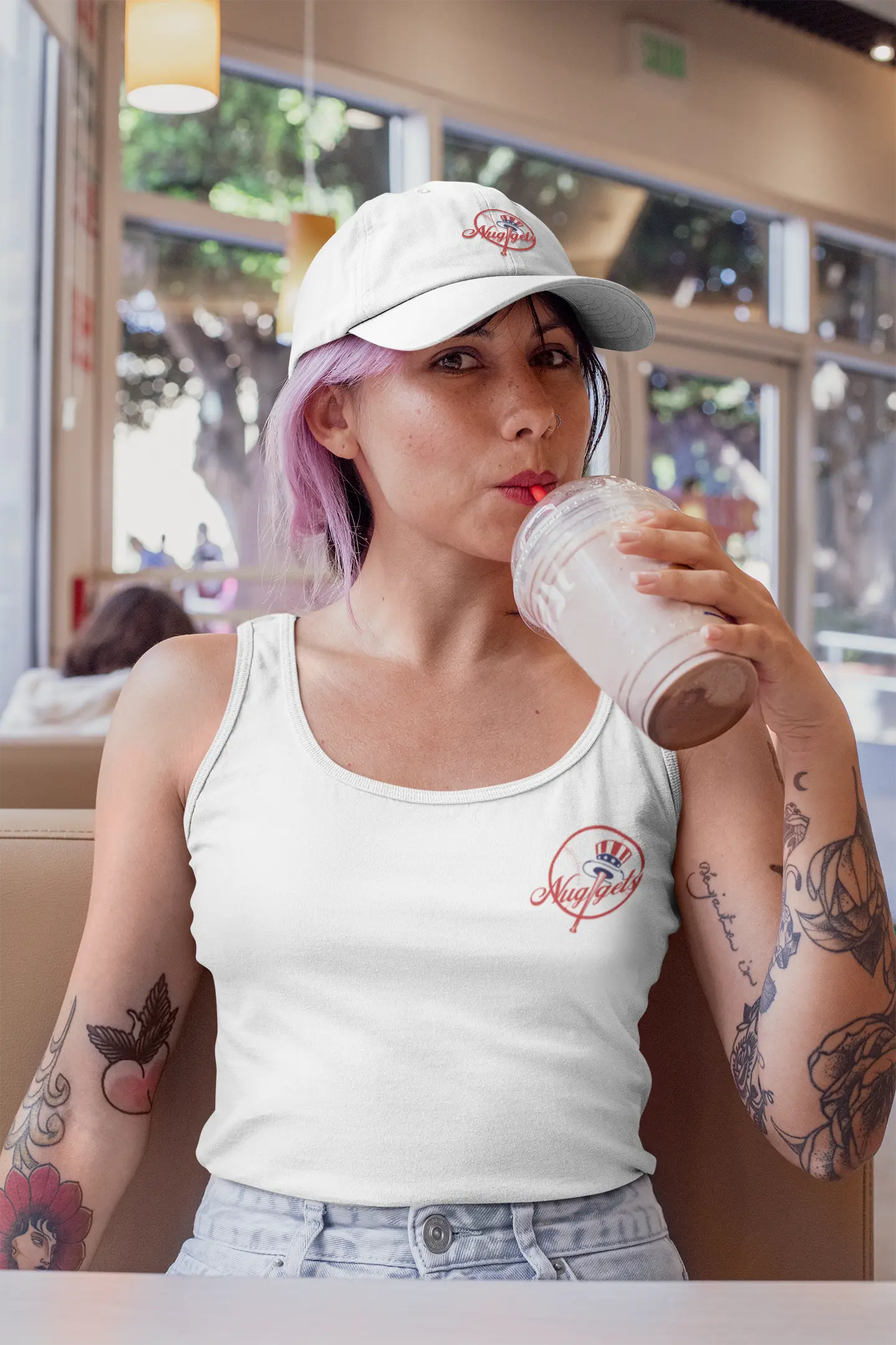 Woman with tattoos wearing white New York Nuggets dad hat and logo tank tops sipping a drink in a cafe.
