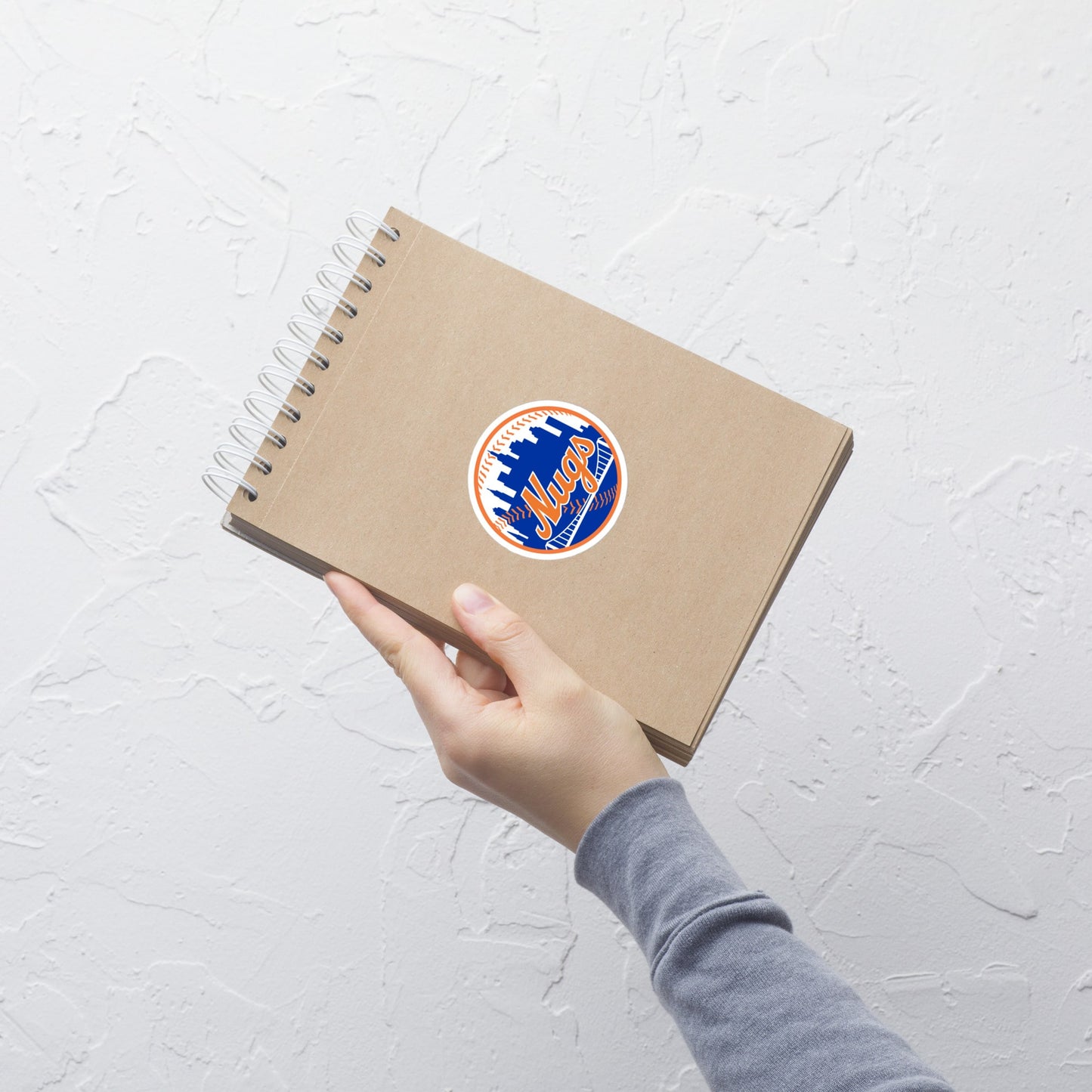 New York Nugs sticker on a brown notebook held in hand against a white textured wall.