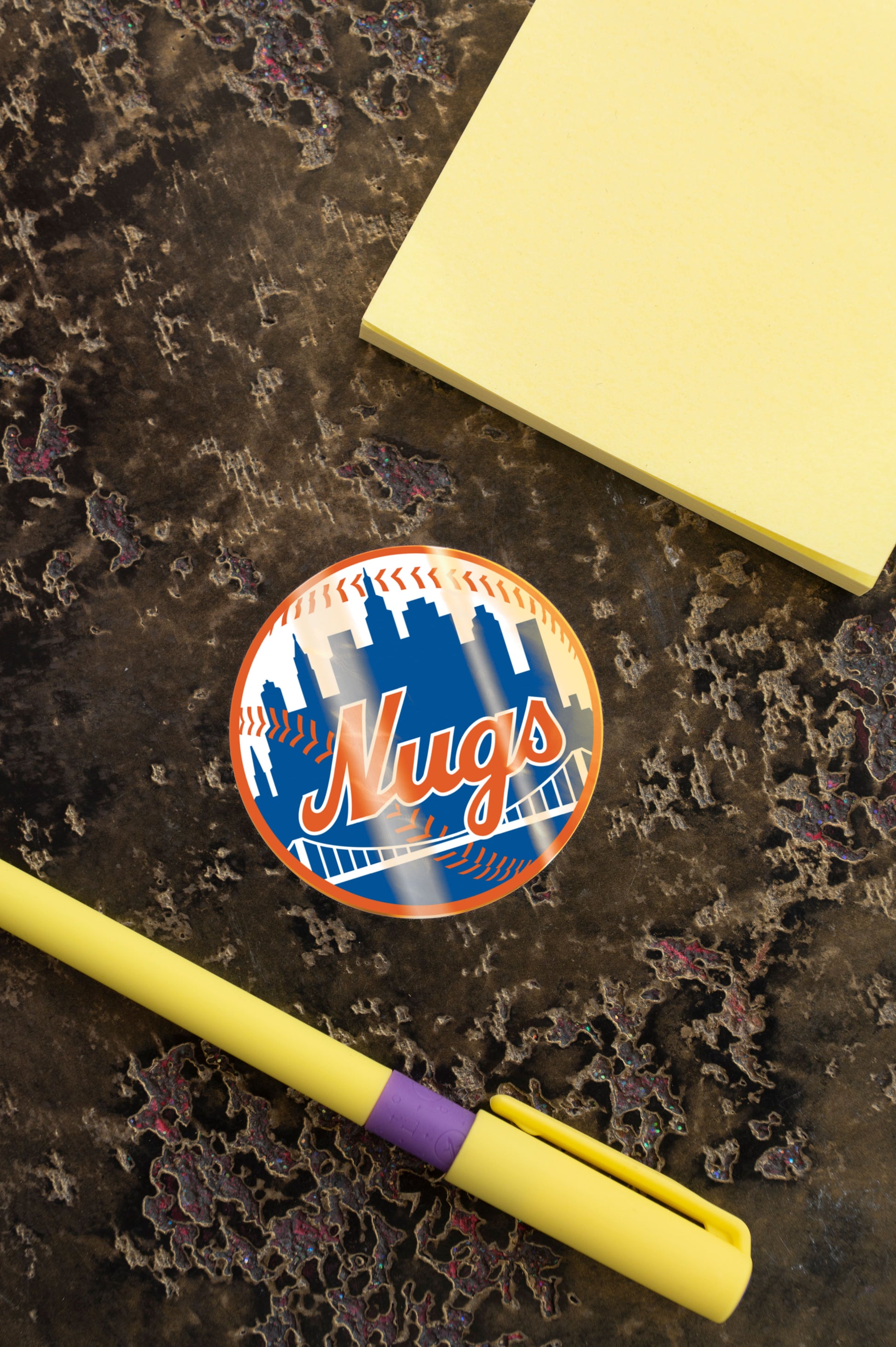 Close-up of New York Nugs sticker beside yellow notepad and pen on a dark marble surface.