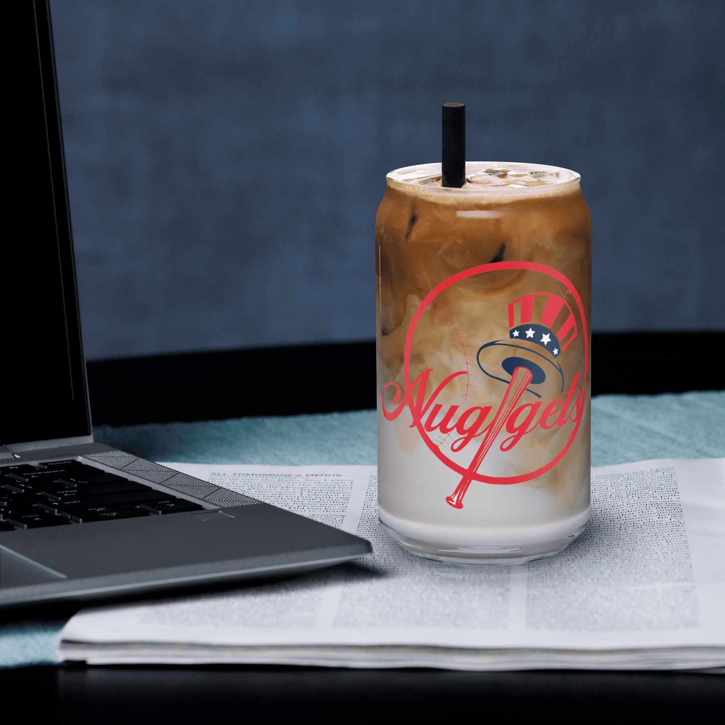 Can-shaped glass with iced latte beside a laptop and newspaper