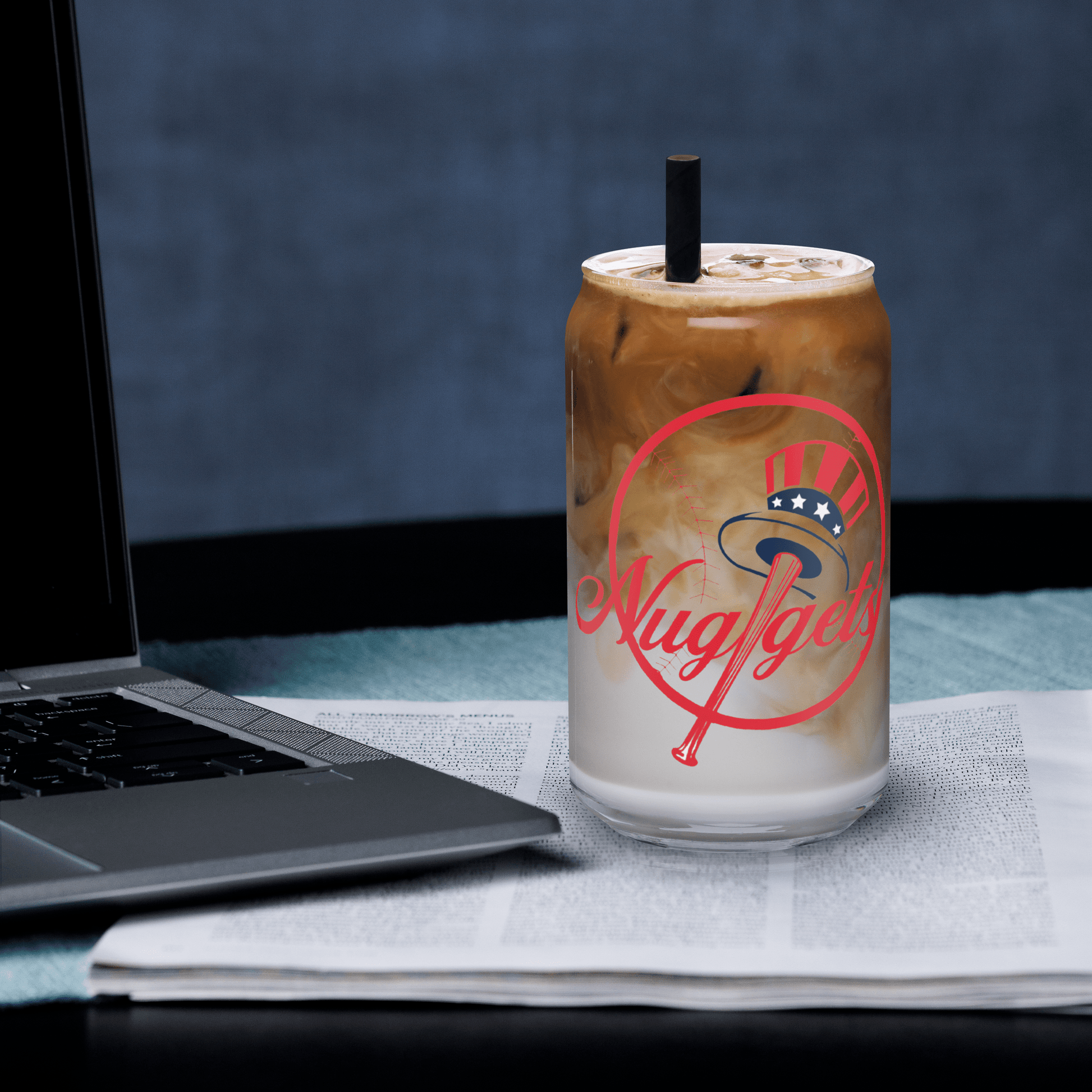Can-shaped glass with iced latte beside a laptop and newspaper