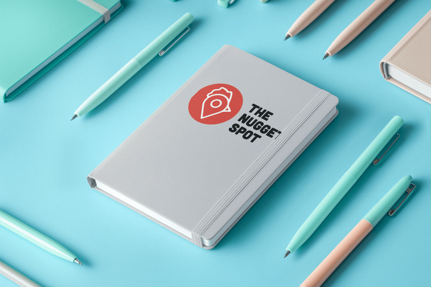 The Nugget Spot hardcover notebook surrounded by pens on a blue background.