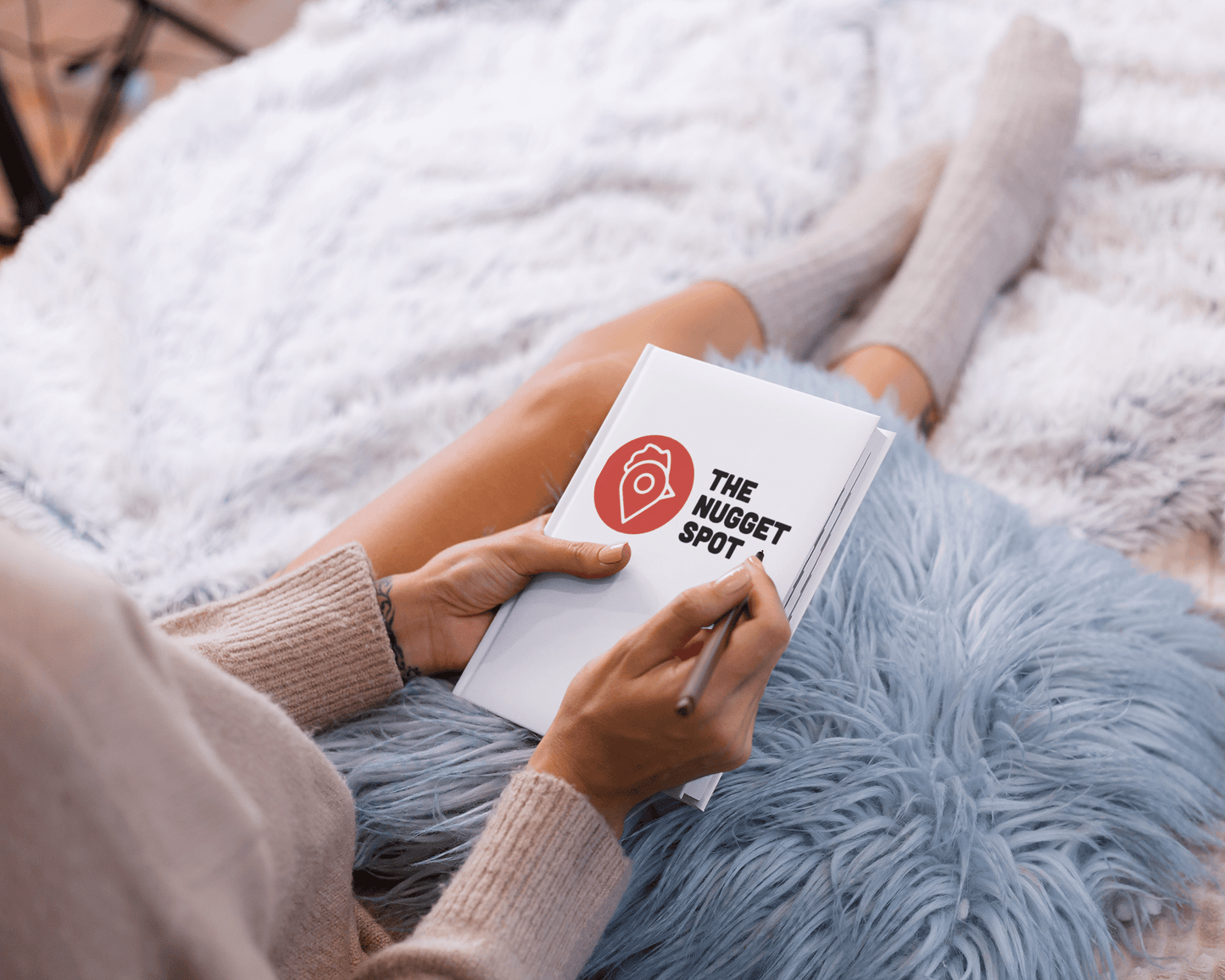 Person writing in The Nugget Spot hardcover notebook on a fluffy blue blanket.