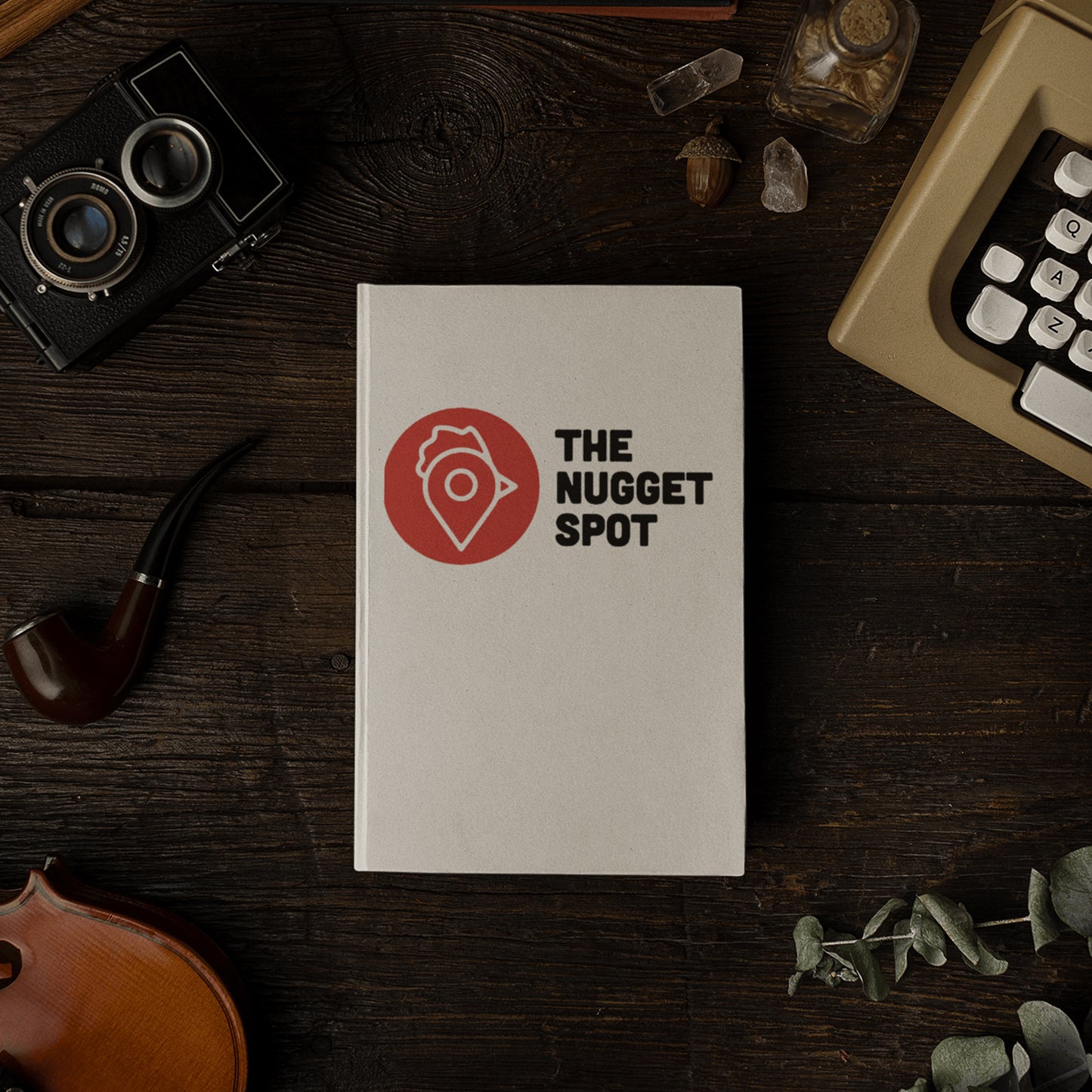 The Nugget Spot hardcover notebook on a rustic wooden desk beside vintage camera and violin.