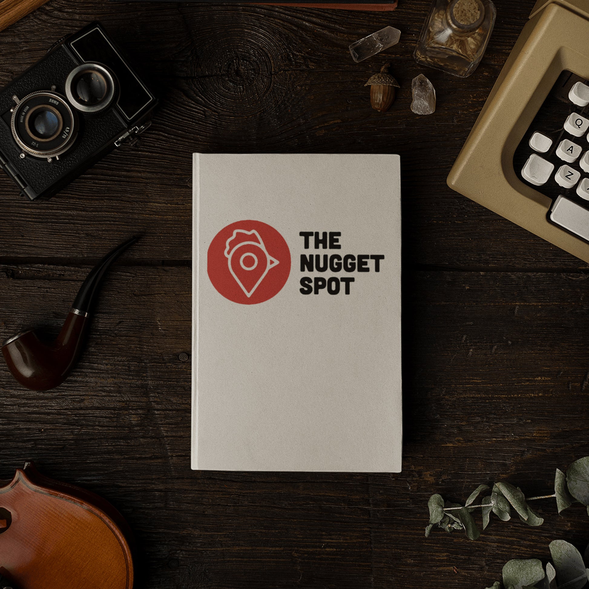 The Nugget Spot hardcover notebook on a rustic wooden desk beside vintage camera and violin.