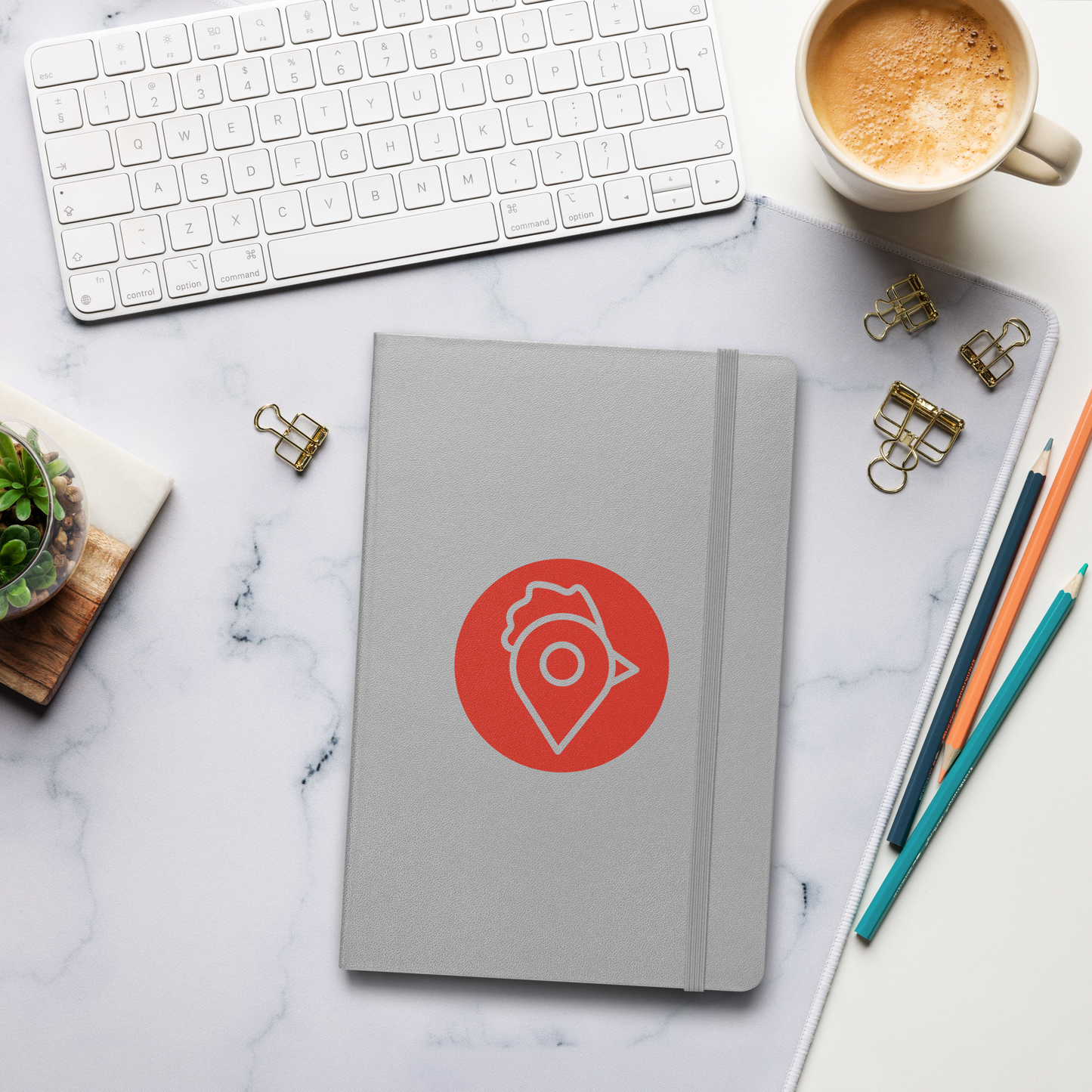 A grey hardcover notebook featuring The Nugget Spot red logo on a marble workspace beside a keyboard and coffee cup
