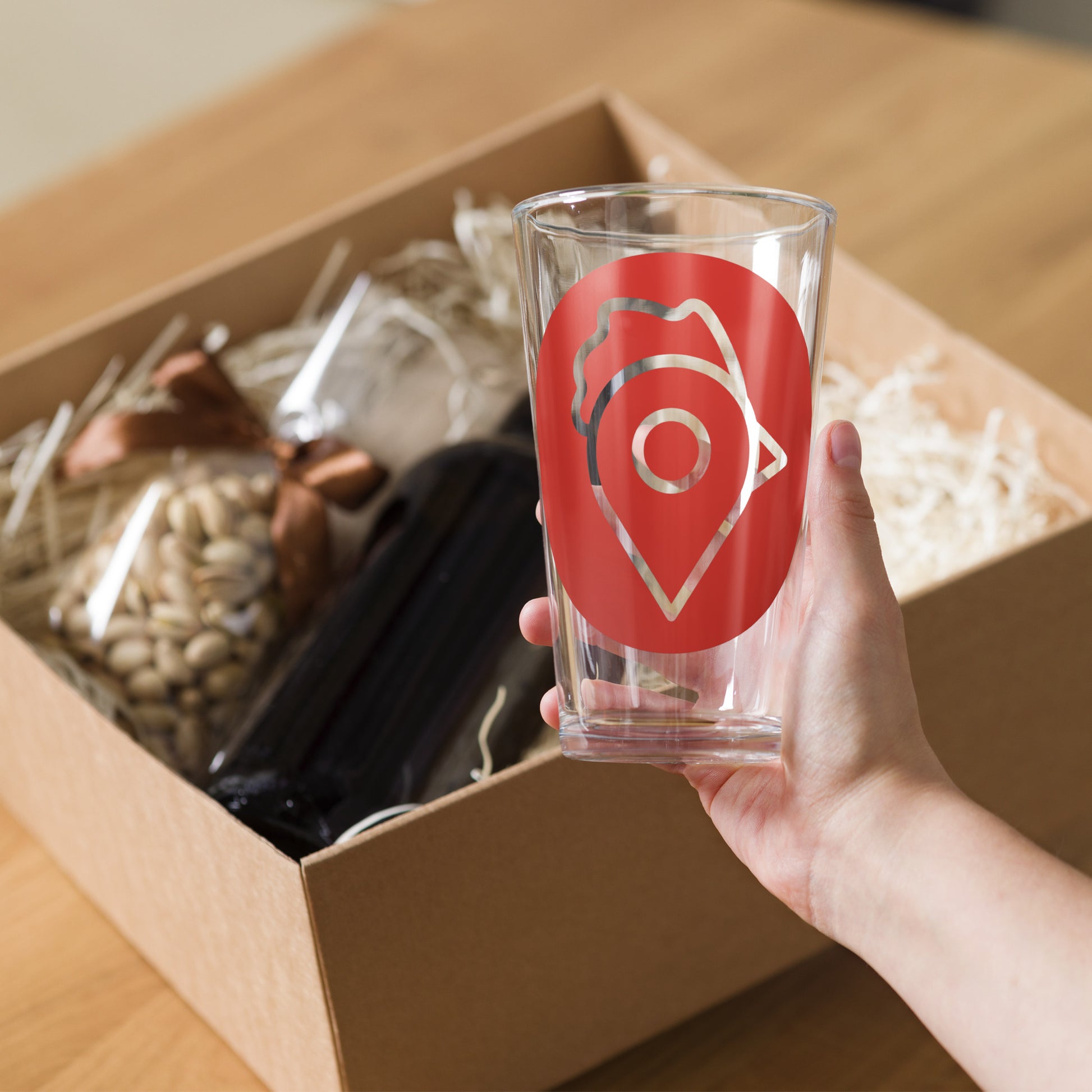 Hand holding The Nugget Spot Signature 16oz Pint Glass beside a gift box filled with treats and a bottle.