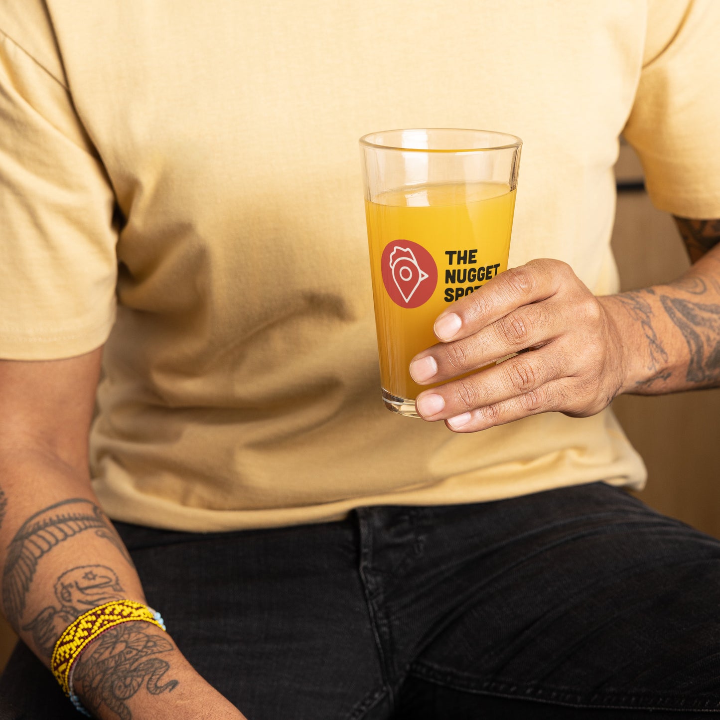 Person holding The Nugget Spot 16oz Pint Glass filled with orange drink – durable UV-printed glassware for everyday use