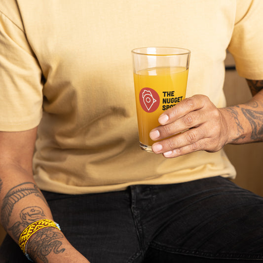 Person holding The Nugget Spot 16oz Pint Glass filled with orange drink – durable UV-printed glassware for everyday use
