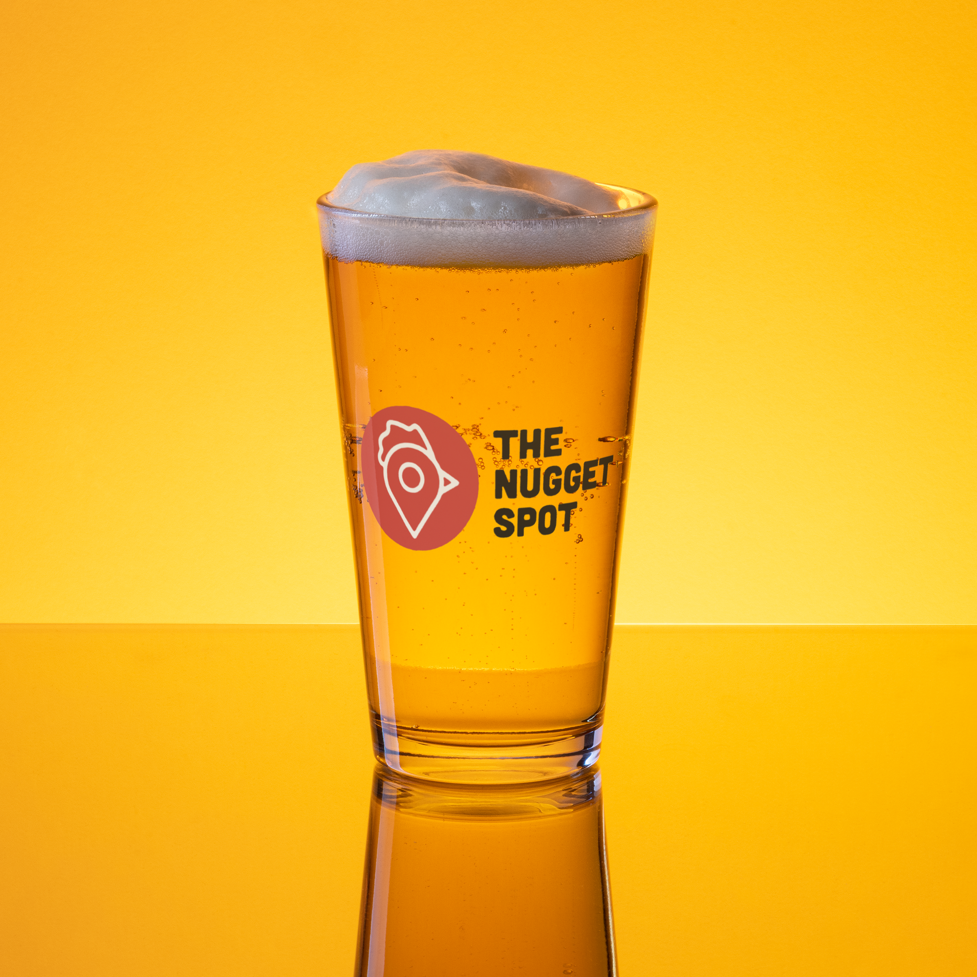 The Nugget Spot 16oz Pint Glass filled with beer and foam on yellow background – vibrant UV-printed logo design