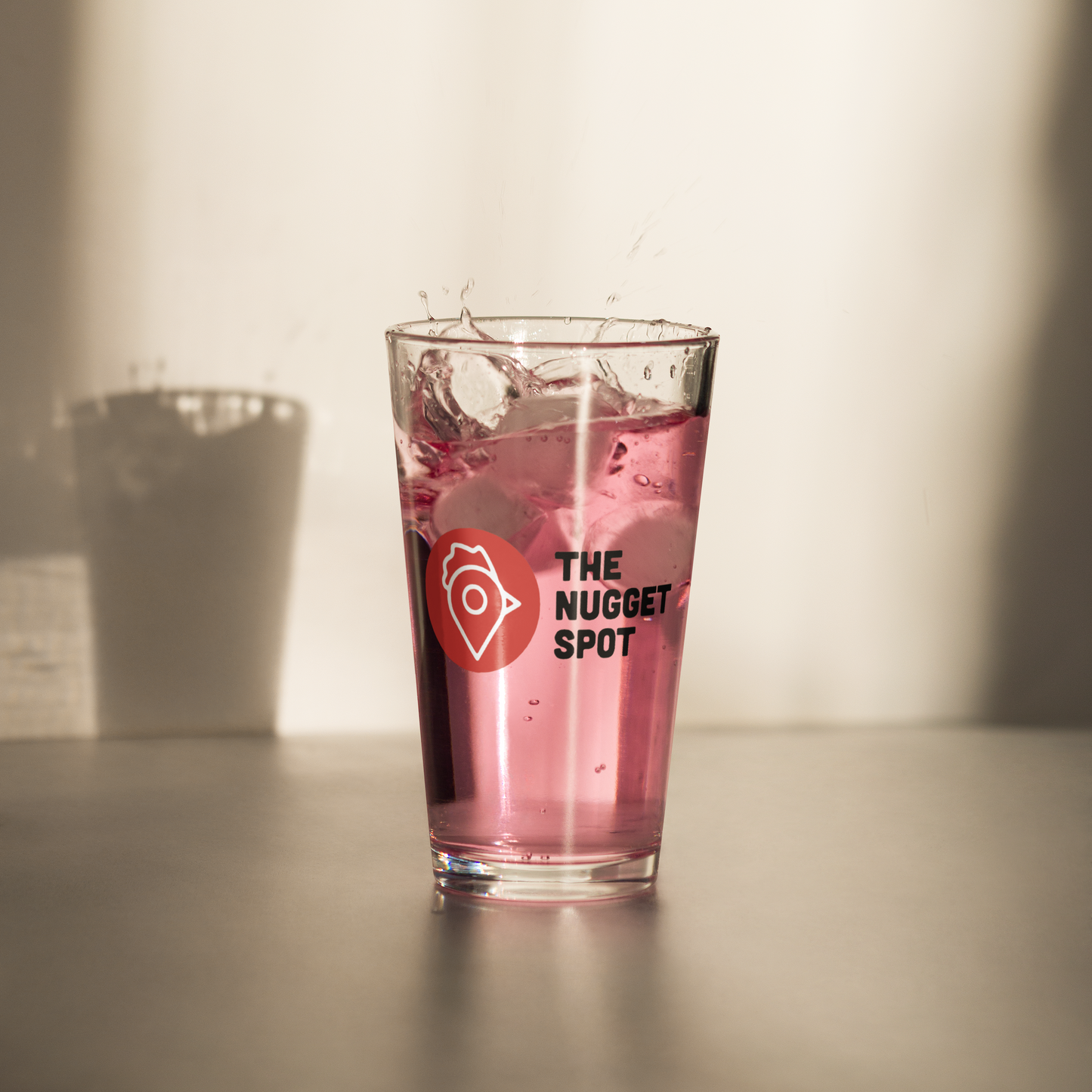 The Nugget Spot 16oz Pint Glass with pink drink splashing – high-quality UV-printed design perfect for cocktails.