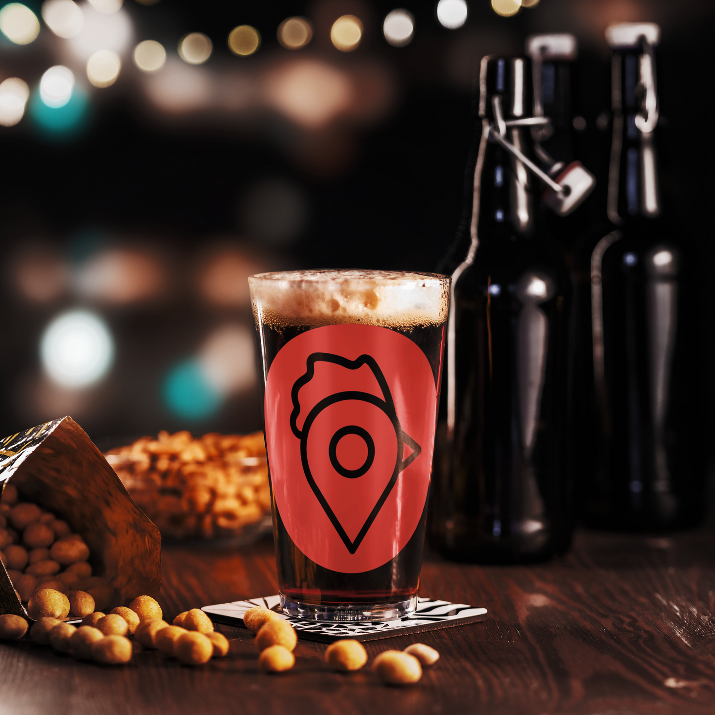 The Nugget Spot Signature 16oz Pint Glass filled with dark beer on a bar table with peanuts and bottles in the background.