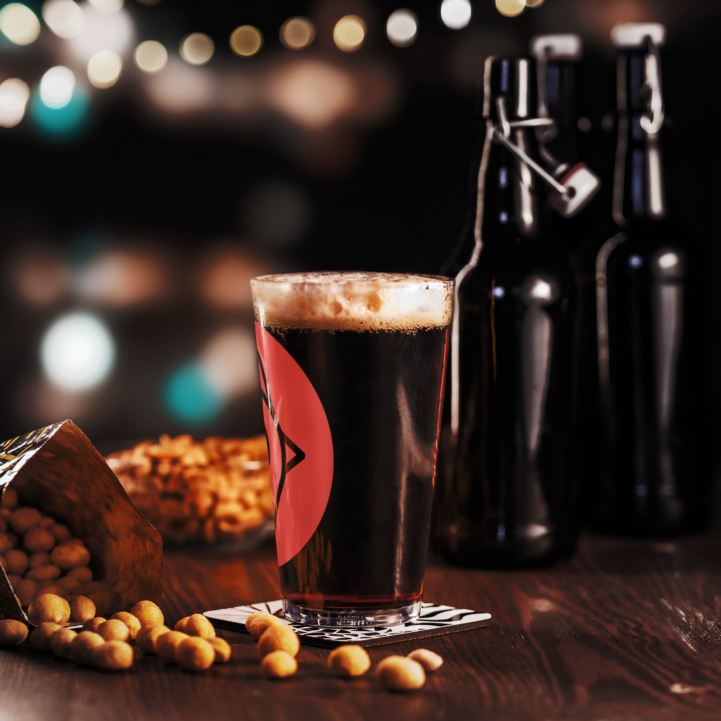 The Nugget Spot Signature 16oz Pint Glass filled with dark beer, surrounded by peanuts and bar decor for a moody night vibe.