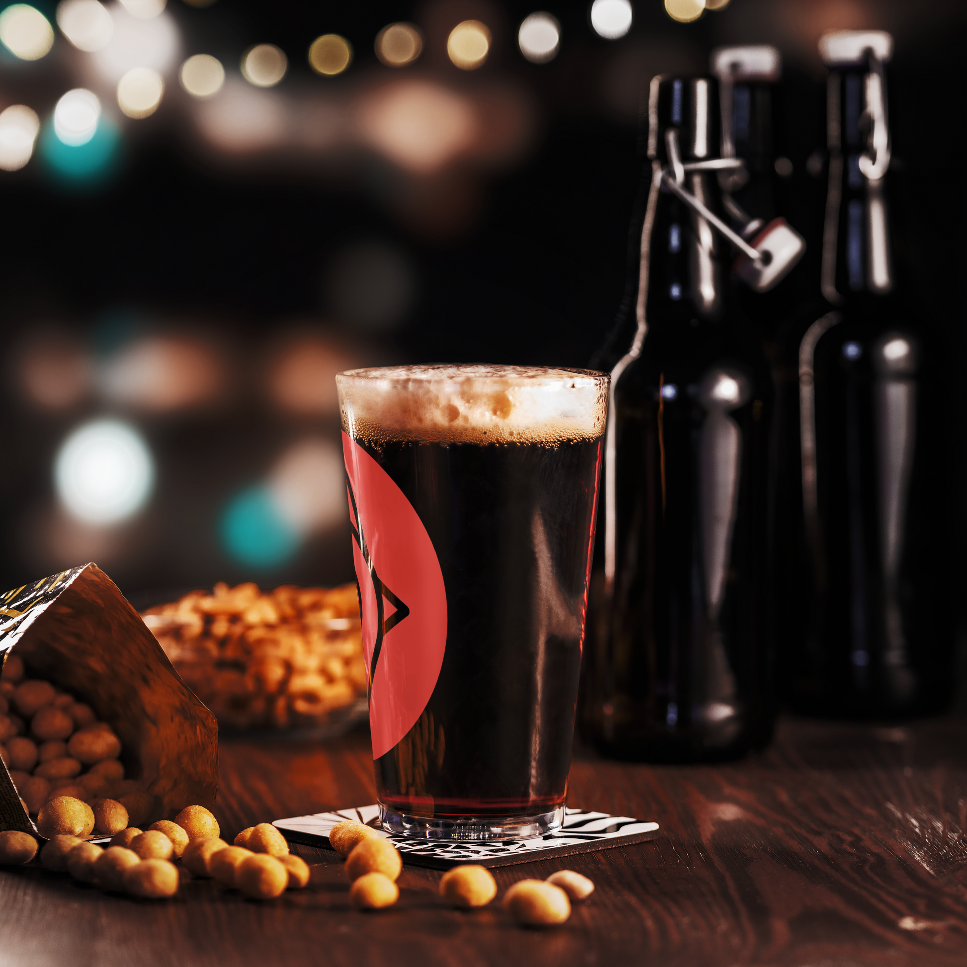 The Nugget Spot Signature 16oz Pint Glass filled with dark beer, surrounded by peanuts and bar decor for a moody night vibe.